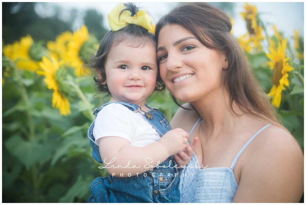 baby with yellow bow and her mom in field of sunflowers