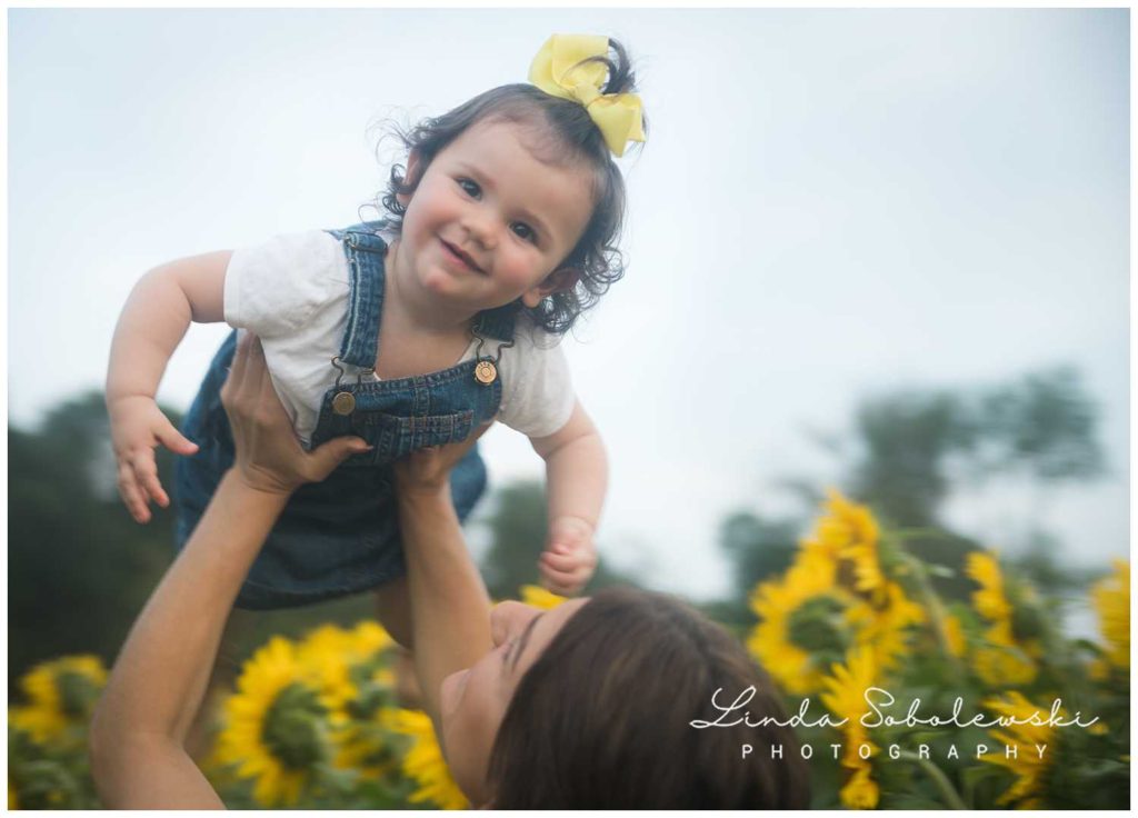 baby with yellow bow being held up over mom's head in field of sunflowers