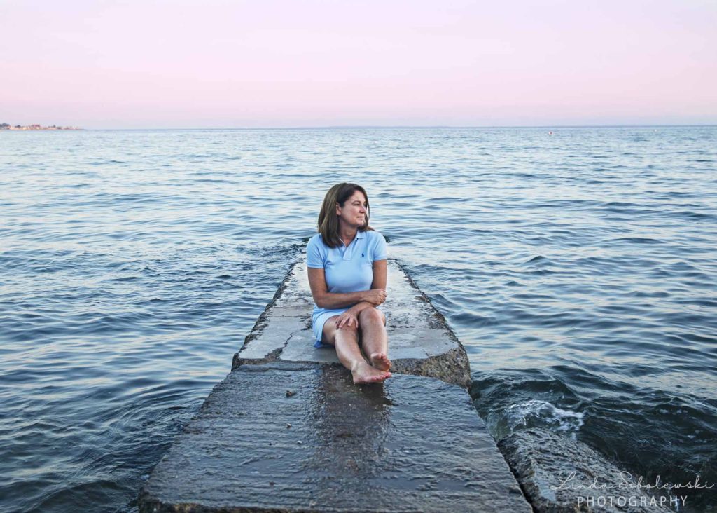 woman in blue dress sitting on a jetty with pink skies behind her, Westbrook lifestyle photographer