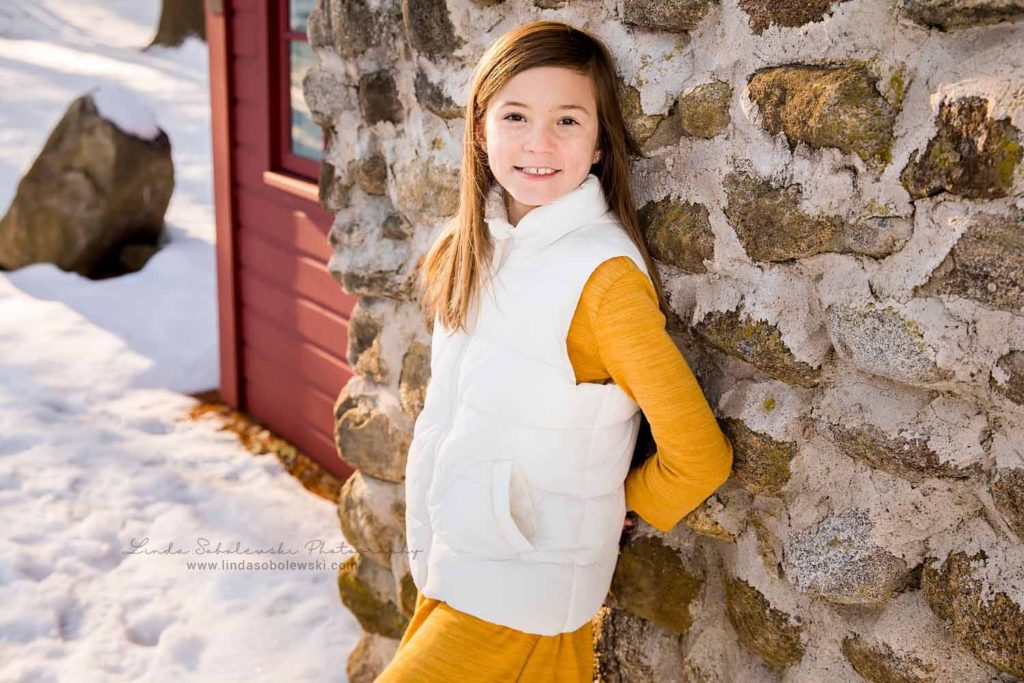 little girl with white vest standing against a stone wall, Family Session in the Snow, Chatfield Hollow Photographer