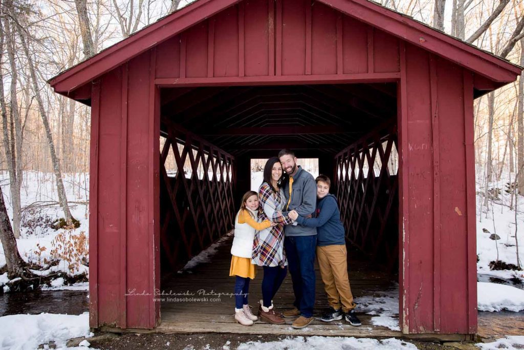 family of four standing together under a red covered bridge, Family Session in the Snow, Old Saybrook Family photographer