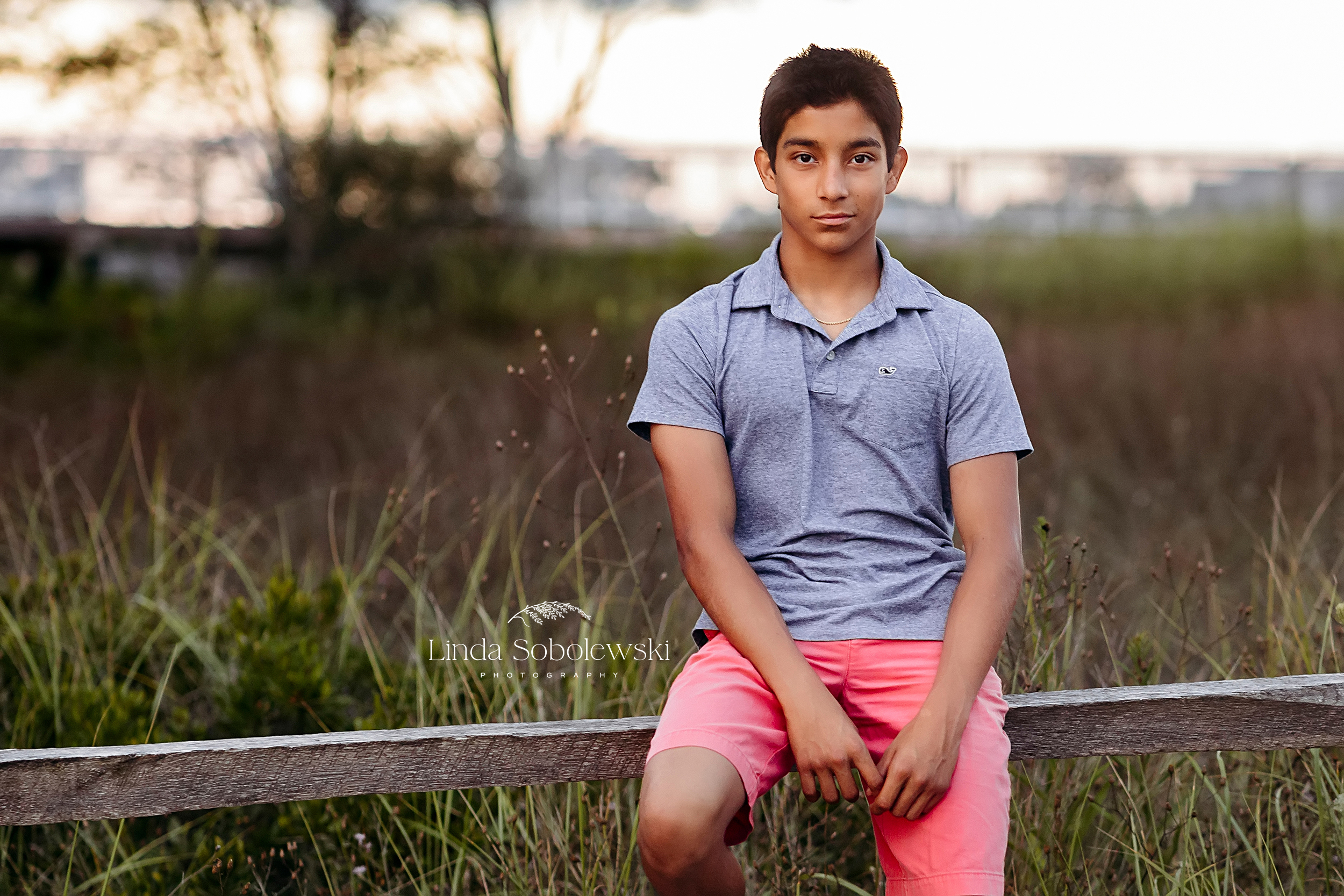 teenage boy in blue shirt leaning against a fence, Senior photos at the beach in Hamonassett beach CT