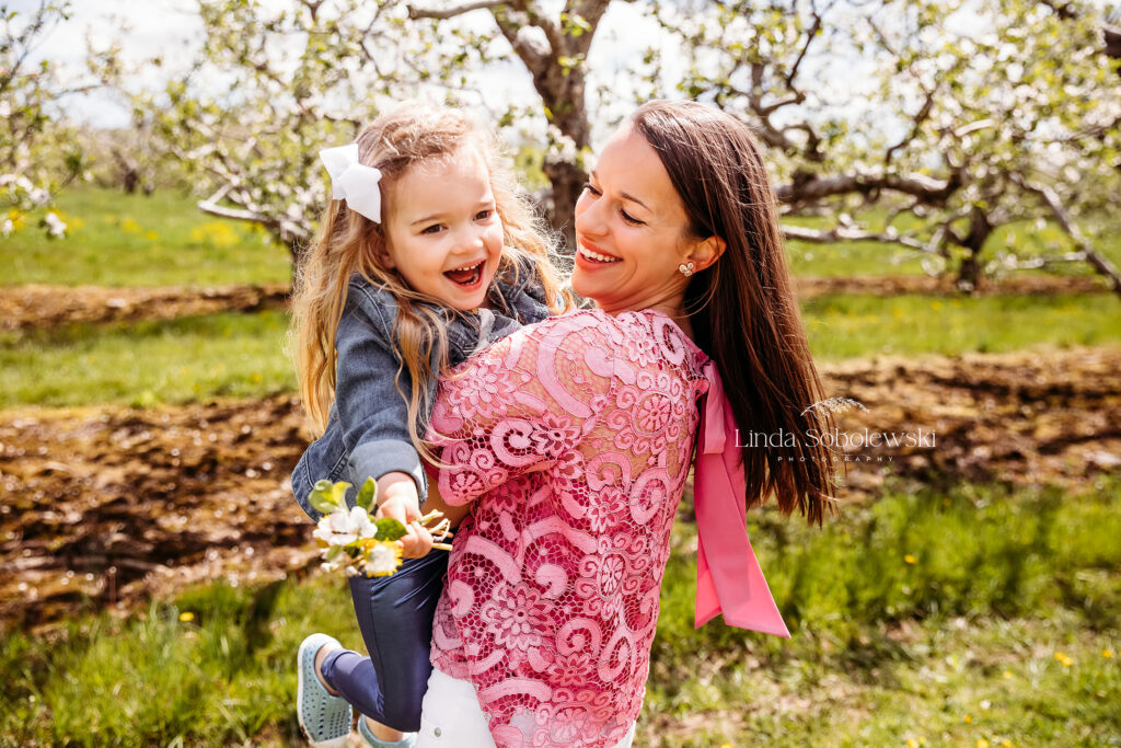 mom in pink shirt twirling her little girl around in an orchard, Best CT Family photographer. Family who is showing us what to wear for their photo session