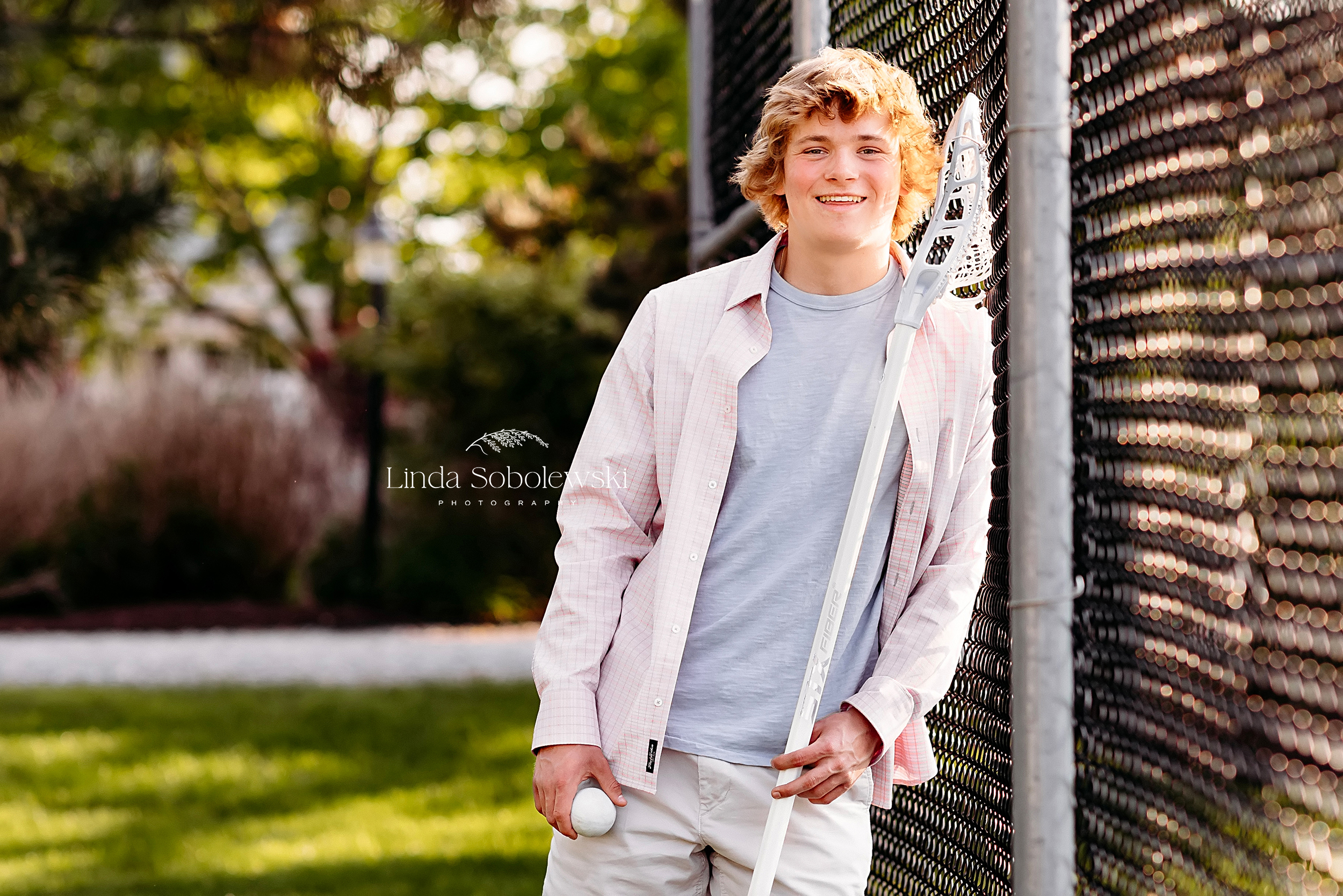 boy in blonde hair and pink shirt holding lacrosse stick for his senior photo session, CT shoreline photographer
