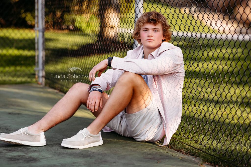 teenage boy in blonde hair sitting against a chain link fence, CT photo session in Old Saybrook, CT