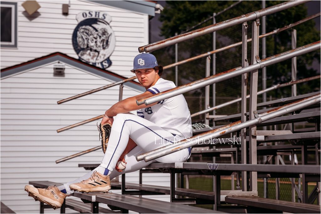 teenage boy in baseball uniform sitting in the metal stands, Old Saybrook CT senior photos and photographer