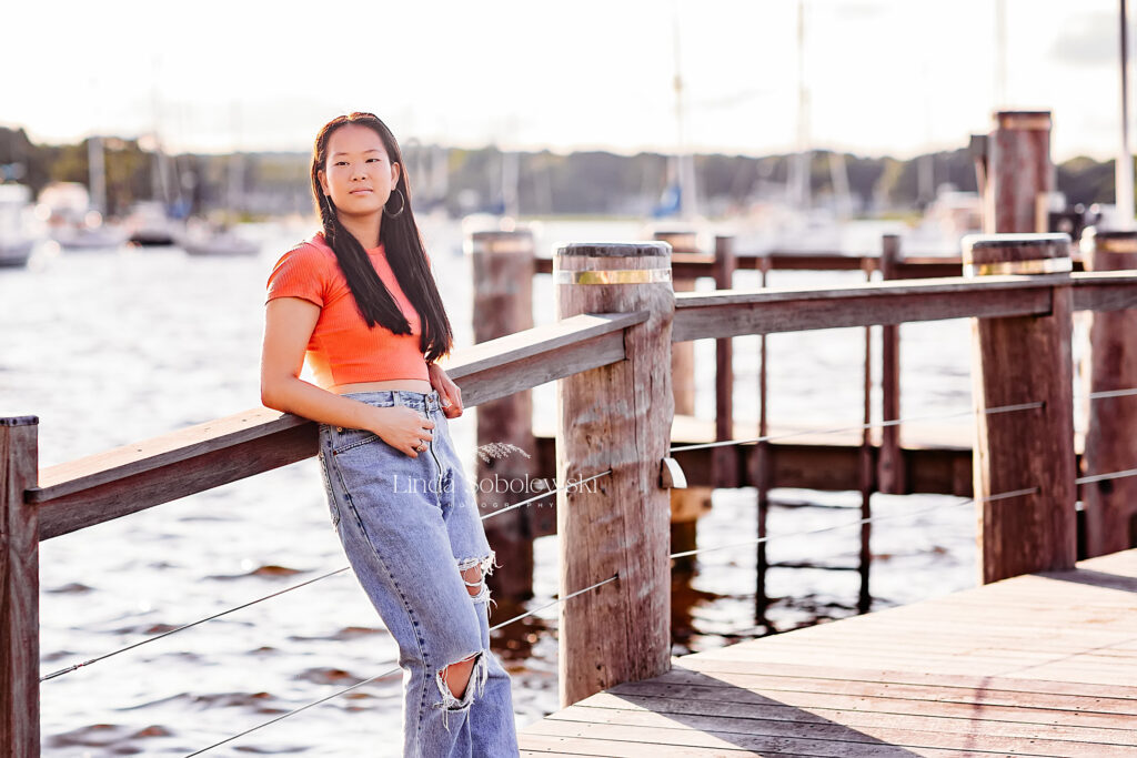 teenage girl in orange shirt and blue jeans leaning against a fence, CT senior photography session