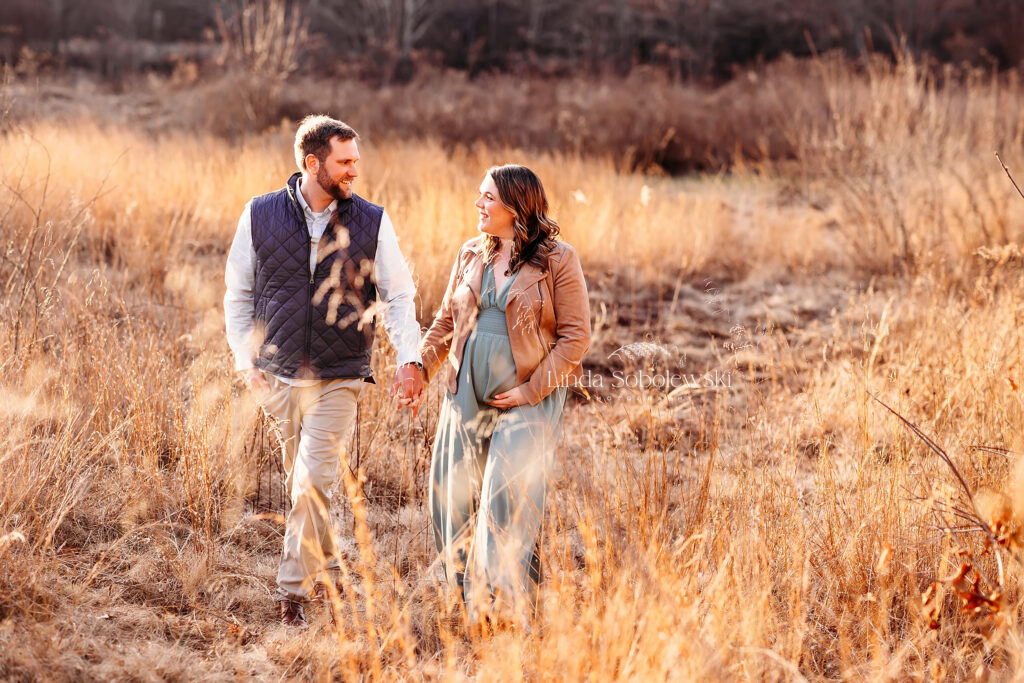 couple holding hands walking in a field, Winter maternity photos in CT