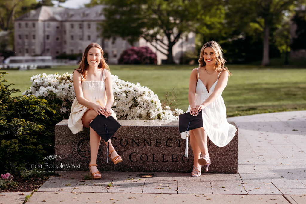 two girls in white dresses holding their graduation caps sitting on a stone wall, CT shoreline senior photographer