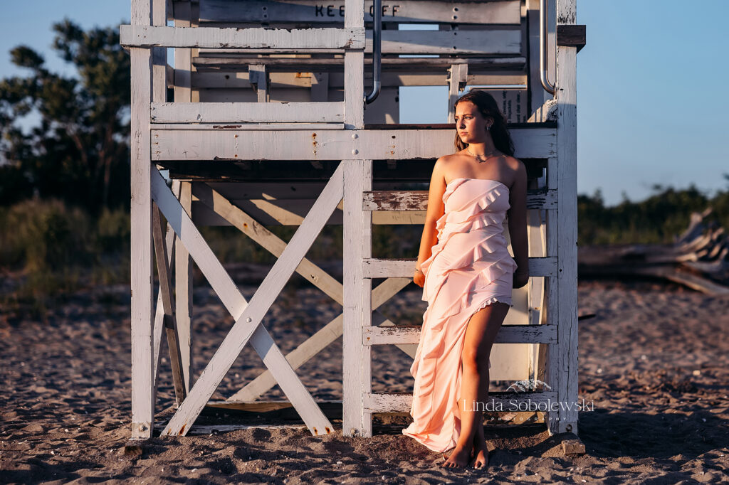 teenage girl in pink dress leaning against a lifeguard chair. Senior photos taken at a park and a beach by CT Senior photographer