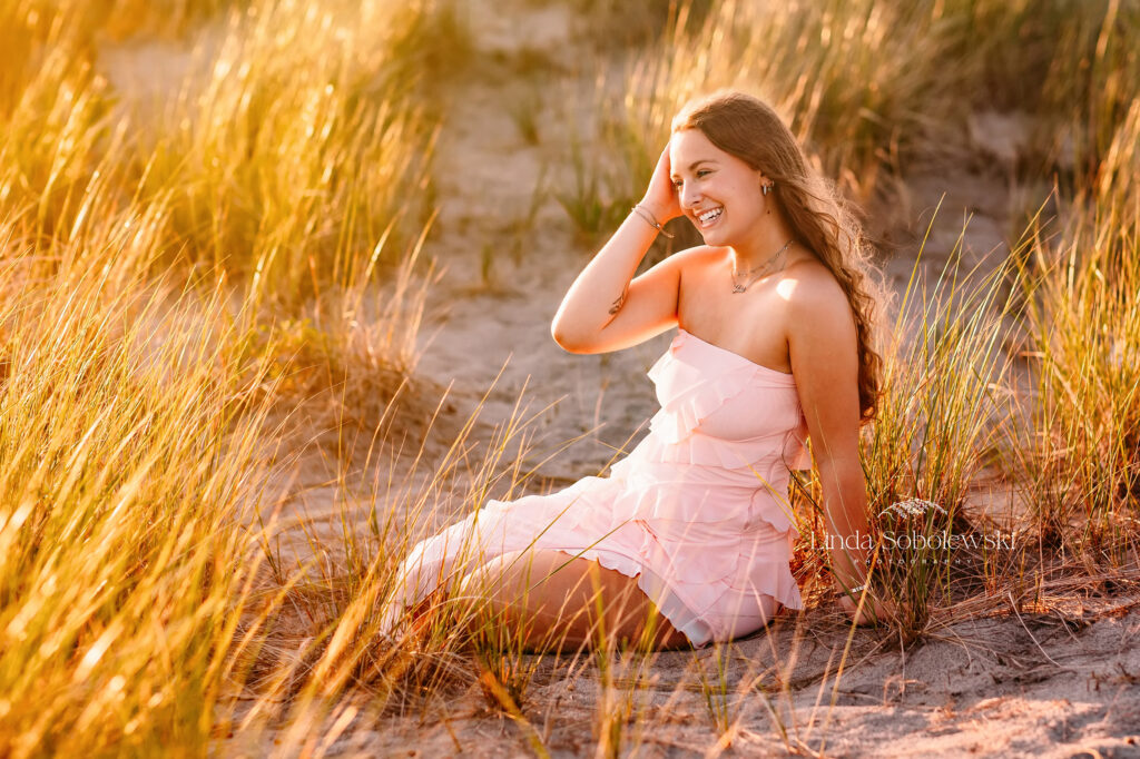 girl in pink dress and long hair sitting in the grass at the beach, Senior photos taken at the beach and park by CT senior photograpehr