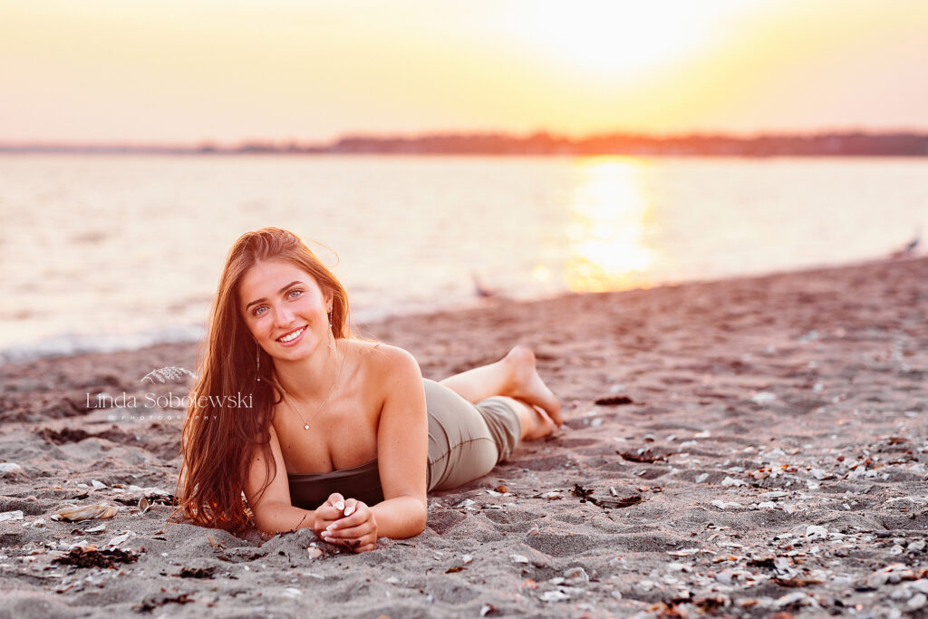 senior girl session at the beach, girl laying down in the sand with the sunset behind her