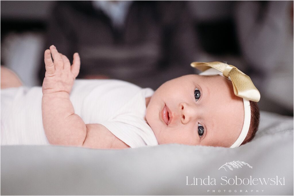 A beautiful newborn baby girl gazes at the camera, wearing a delicate golden bow during her in-home newborn session.