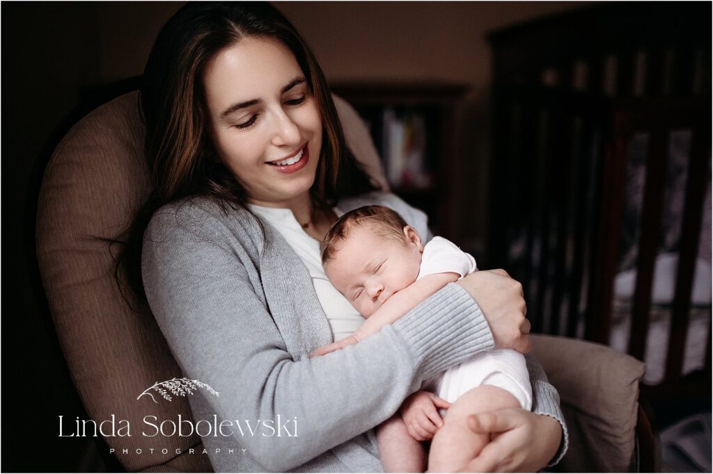 A mother smiles while cradling her peacefully sleeping newborn during an in-home newborn session.