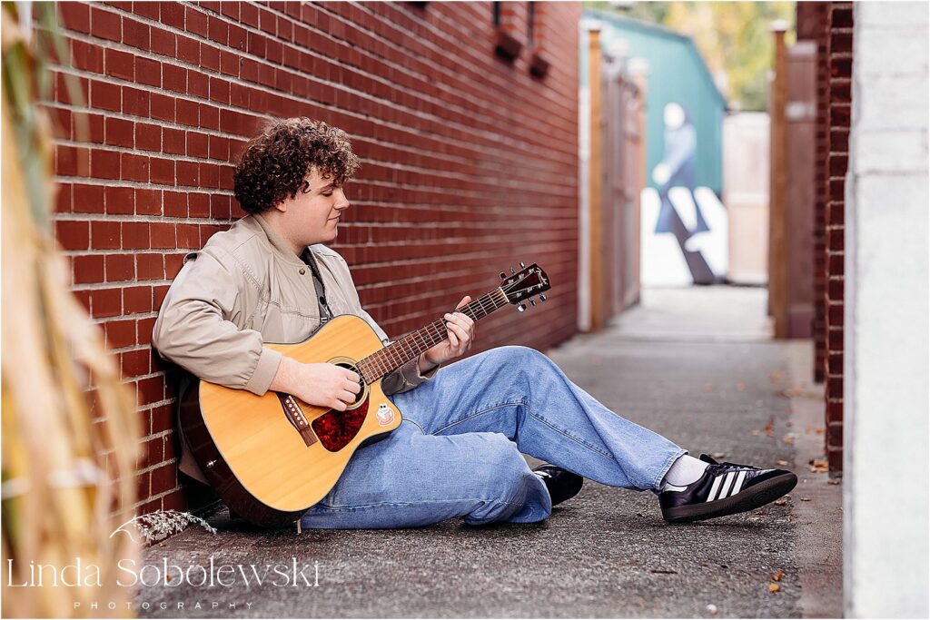 Senior guy leans against a brick wall playing his acoustic guitar in a unique urban setting for his senior photography session in Old Saybrook, CT.