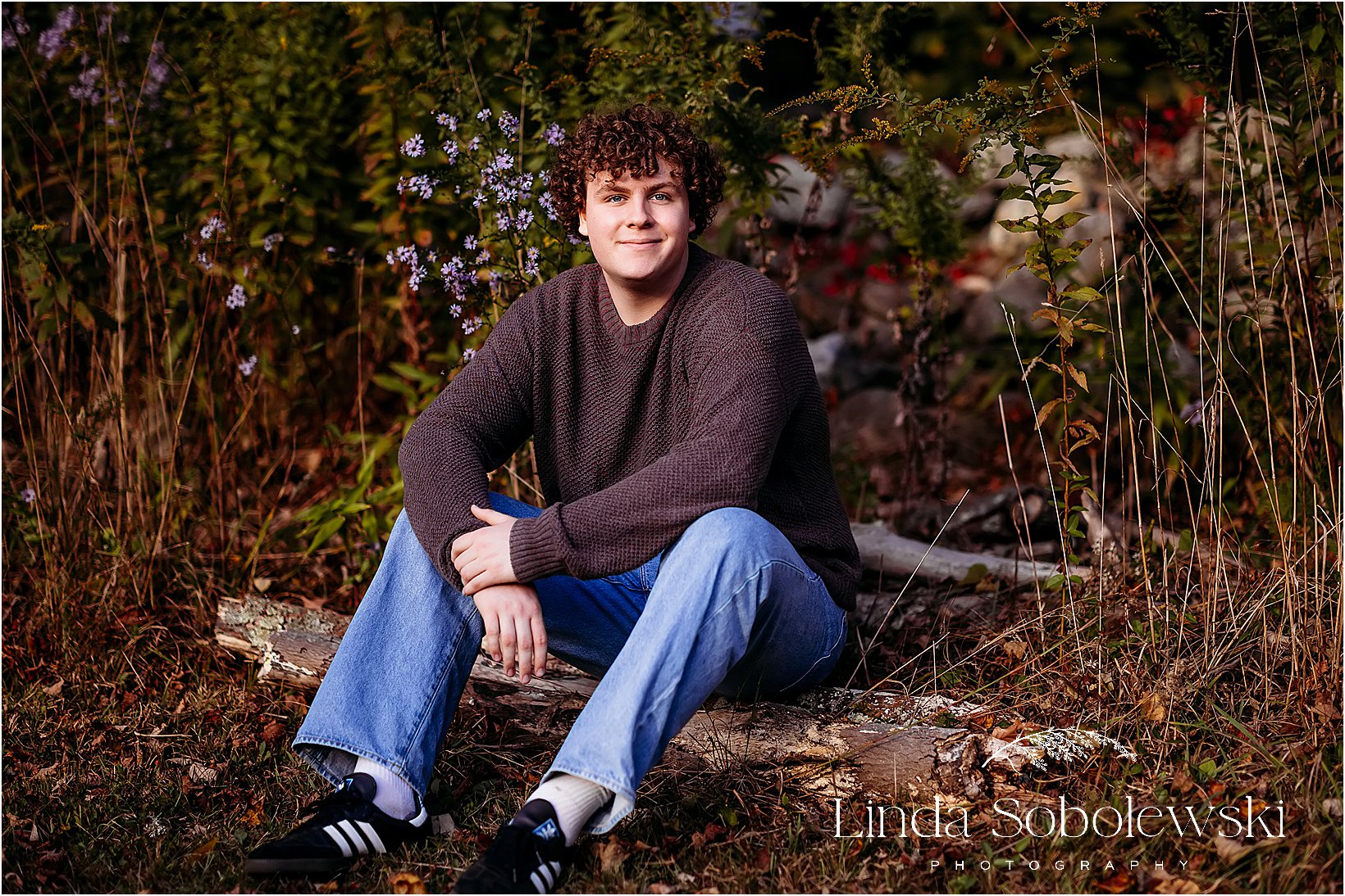 Senior portrait of a young man sitting on a rustic log, surrounded by wildflowers and natural textures in Old Saybrook, CT.