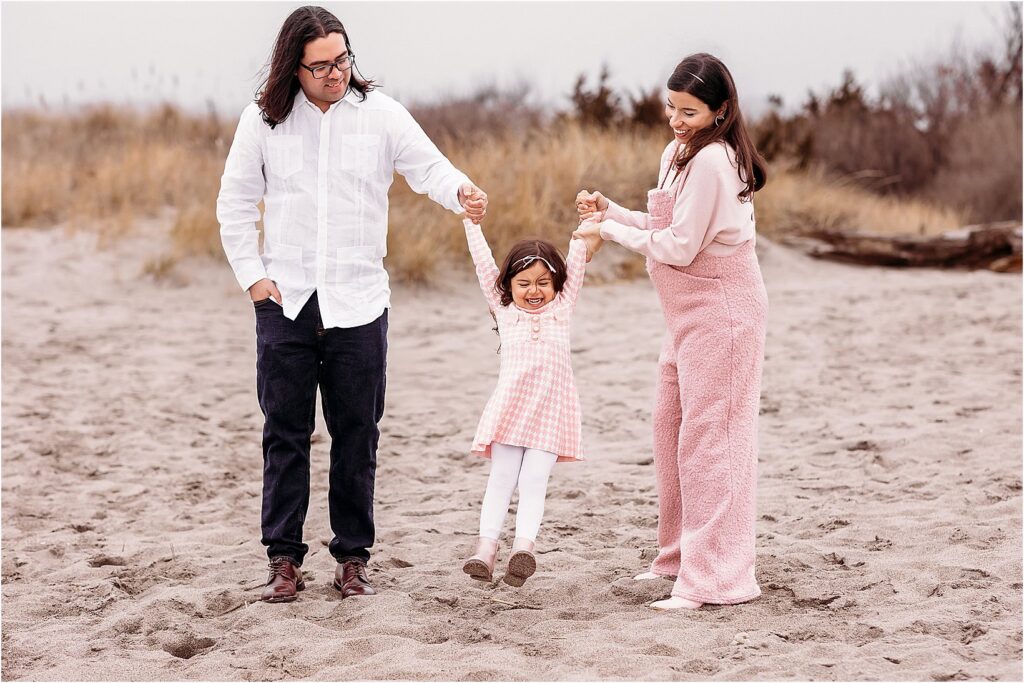 Little girl being swung by her parents during a joyful maternity beach session in Connecticut.