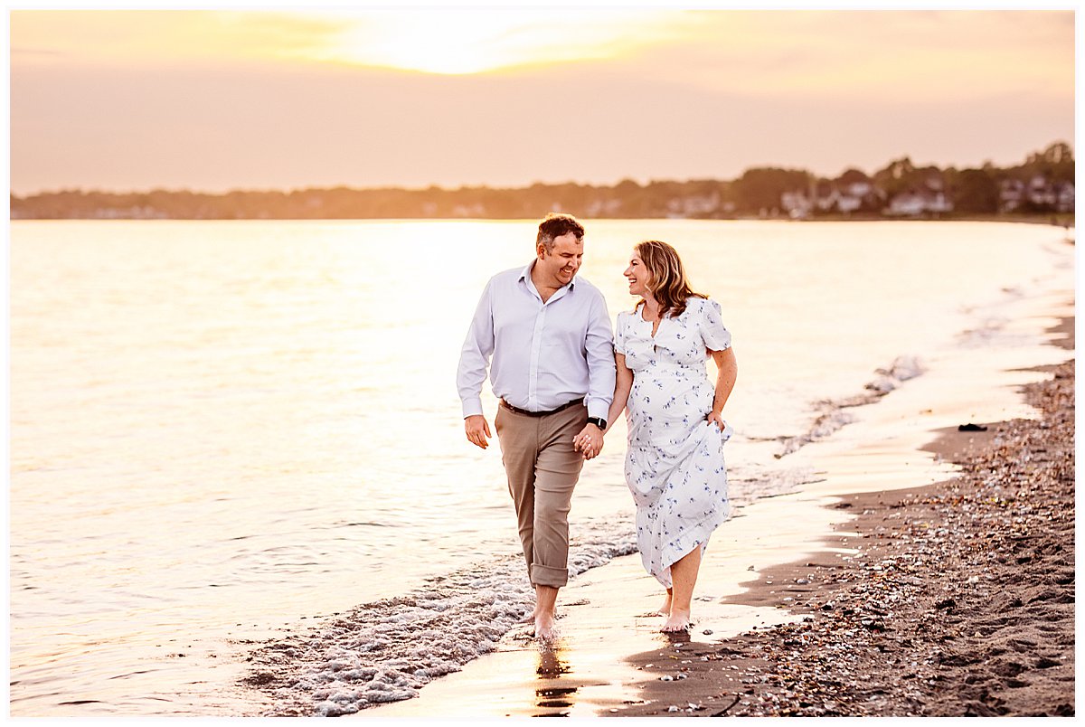 Maternity photos at the beach in CT with an expecting couple walking the shoreline at sunset at Hammonasset