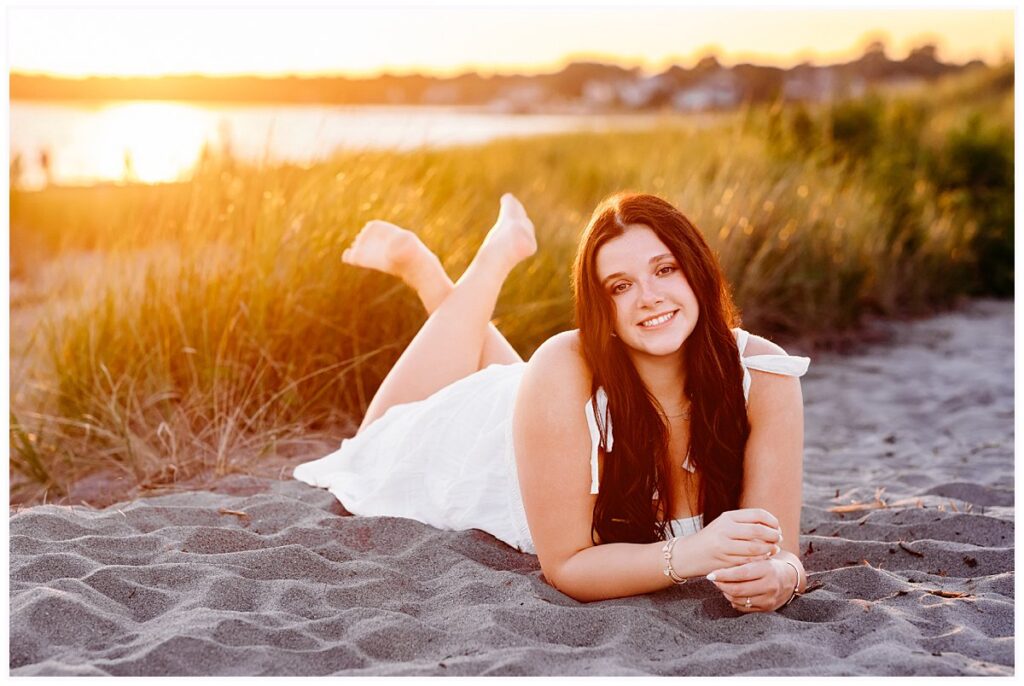 teenage girl in white dress laying in the sand as the sun sets behind her. CT shoreline senior Pictures