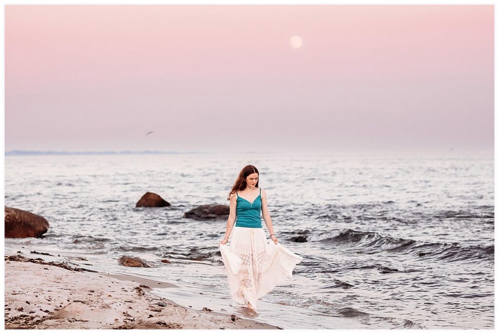 Park for her Senior Pictures in Connecticut. Emma walks along the beach at Harkness Memorial State Park beneath a soft pink sky and moon during her Senior Pictures in Connecticu