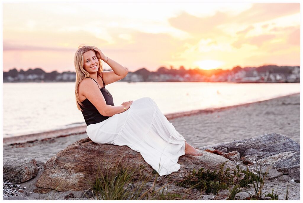 Old Saybrook Senior Session on a Boat featuring Elle sitting on shoreline rocks with a colorful summer sunset behind her.