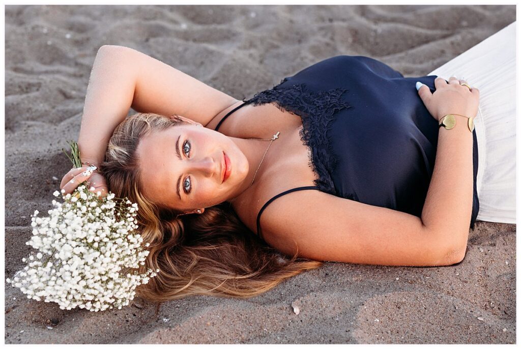Close-up senior portrait of Elle holding white flowers while lying on the sand at an Old Saybrook beac